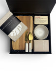 Premium corporate gift box containing hand-printed linen tea towel, handcrafted ceramic dishes, artisan soap, ecosoy candle, wood serving board and marble-handled cheese knives, styled on a neutral background