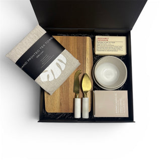 Premium corporate gift box containing hand-printed linen tea towel, handcrafted ceramic dishes, artisan soap, ecosoy candle, wood serving board and marble-handled cheese knives, styled on a neutral background
